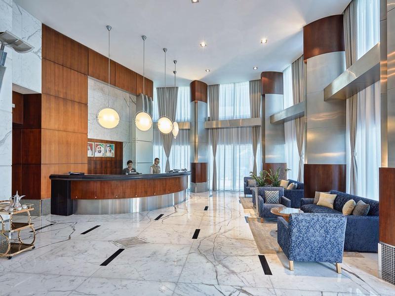 Jasna lobby hotelowa z marmurową podłogą, drewnianymi panelami, recepcją i przytulnymi miejscami do siedzenia.