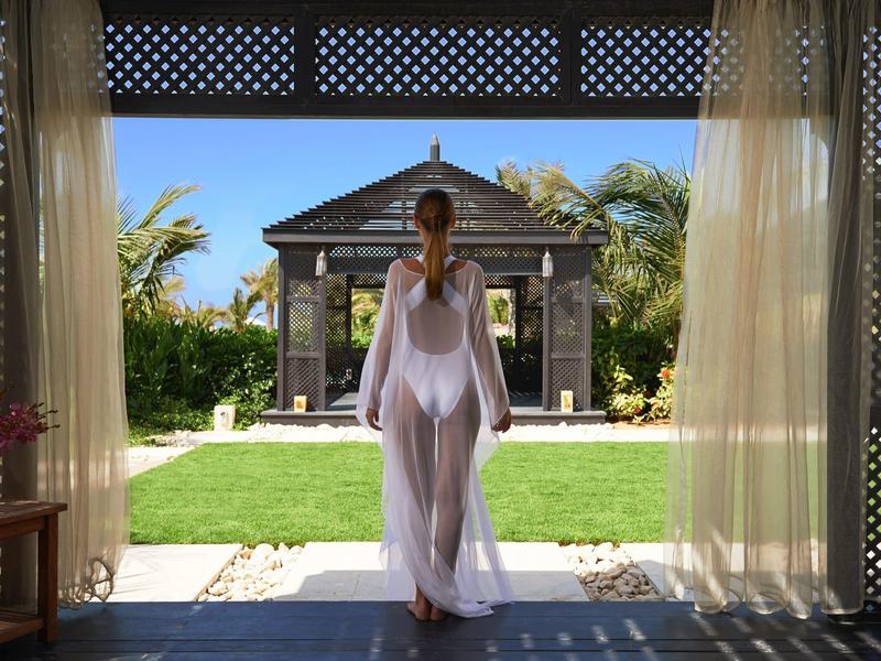 Frau in weißem Kleid steht in einer offenen Tür mit Blick auf Garten und Pavillon.