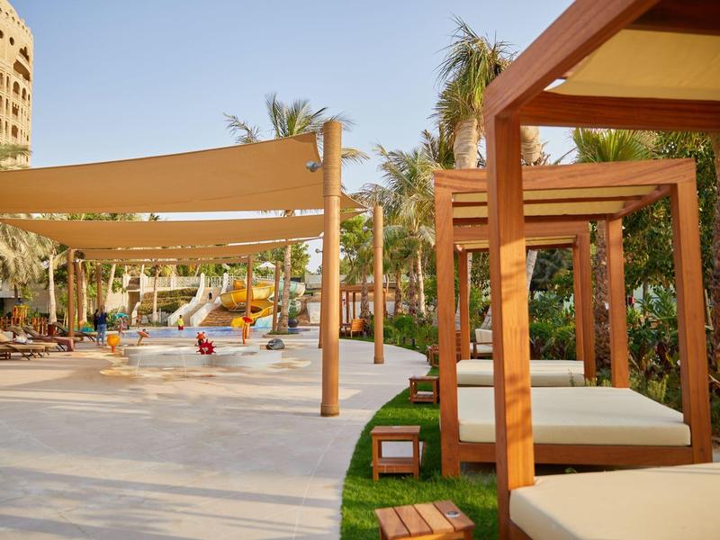 Hotelgarten mit Liegen unter Holzpergolen und Kinderspielplatz im Hintergrund bei klarem Himmel