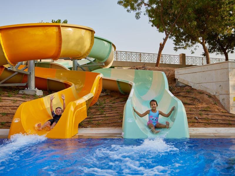 Zwei Personen rutschen auf bunten Wasserrutschen in einen Pool bei sonnigem Wetter.