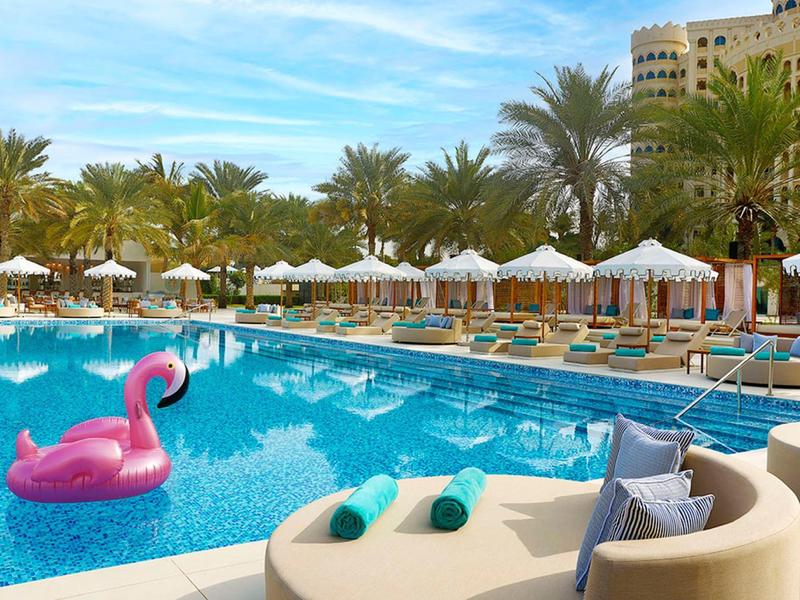 Großer Hotelpool mit Liegestühlen, weißen Sonnenschirmen und Palmen unter blauem Himmel.