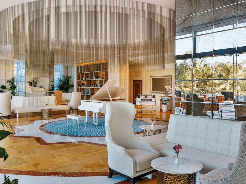 Eleganter Hotel-Loungebereich mit weißen Sesseln, Klavier und großen Fenstern mit Blick auf Palmen.