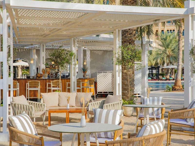 Helle Outdoor-Lounge mit Stühlen, Tischen und Pflanzen unter einem überdachten Bereich.