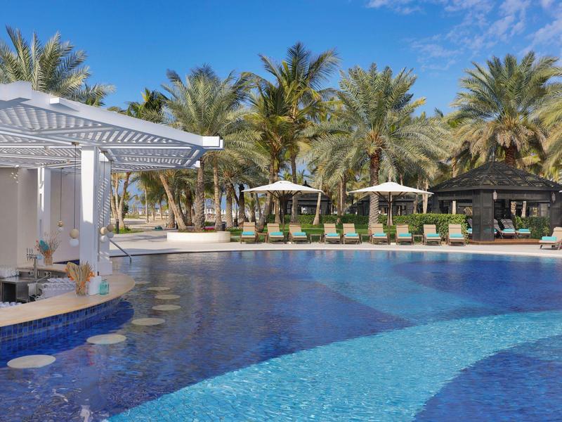 Großer Resortpool mit Sonnenliegen und Palmen unter blauem Himmel.