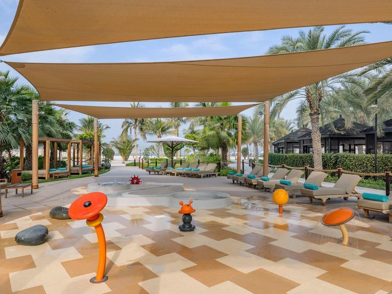 Kinderbereich mit Wasserspielplatz, schattigen Segeln, Sitzgelegenheiten und Palmen im Hintergrund.