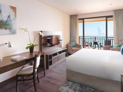 Helles Hotelzimmer mit großem Bett, Schreibtisch, Fernseher und Balkon mit Meerblick.