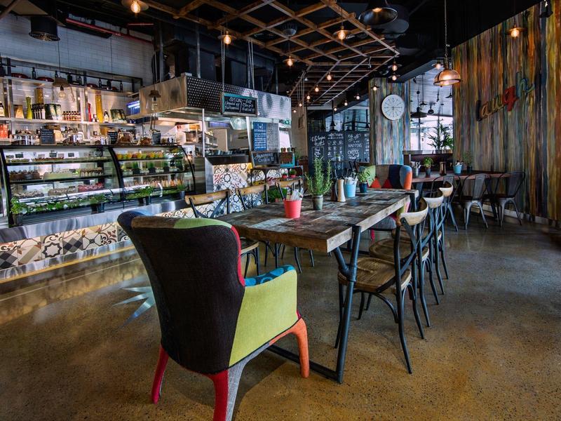 Modern eingerichtetes Café mit bunten Stühlen, langen Holztischen und einer großen Theke.