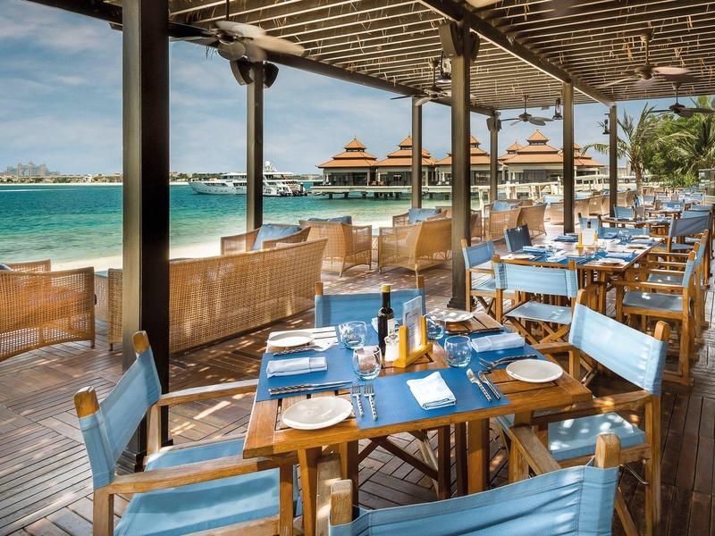 Offenes Restaurant am Meer mit Holztischen, blauen Stühlen und Blick auf Palmen und Wasser.
