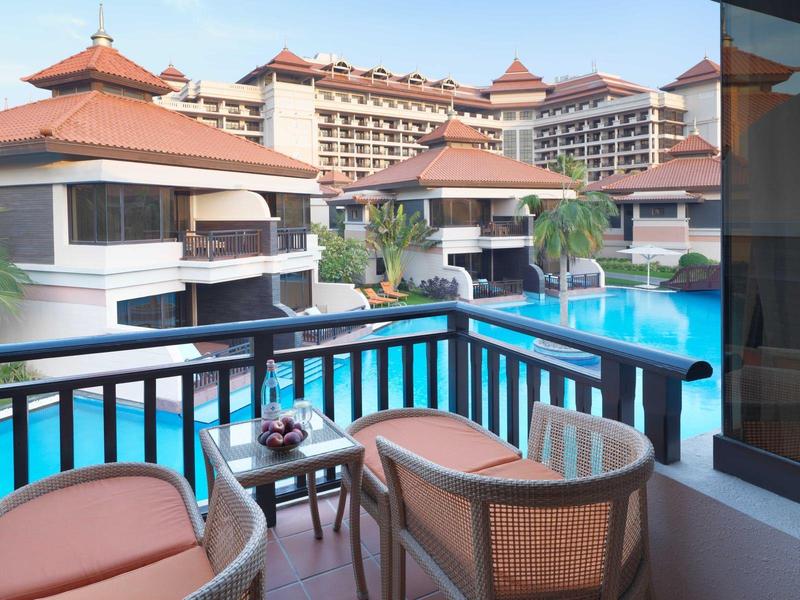 Balkon mit Stühlen und Tisch, Blick auf Pool und Hotelgebäude mit roten Dächern bei Sonnenschein.