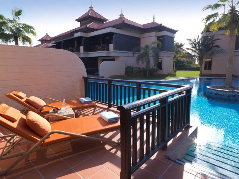 Zwei orange Liegestühle auf Terrasse neben blauem Pool und mehrstöckigem Haus mit Palmen im Hintergrund.