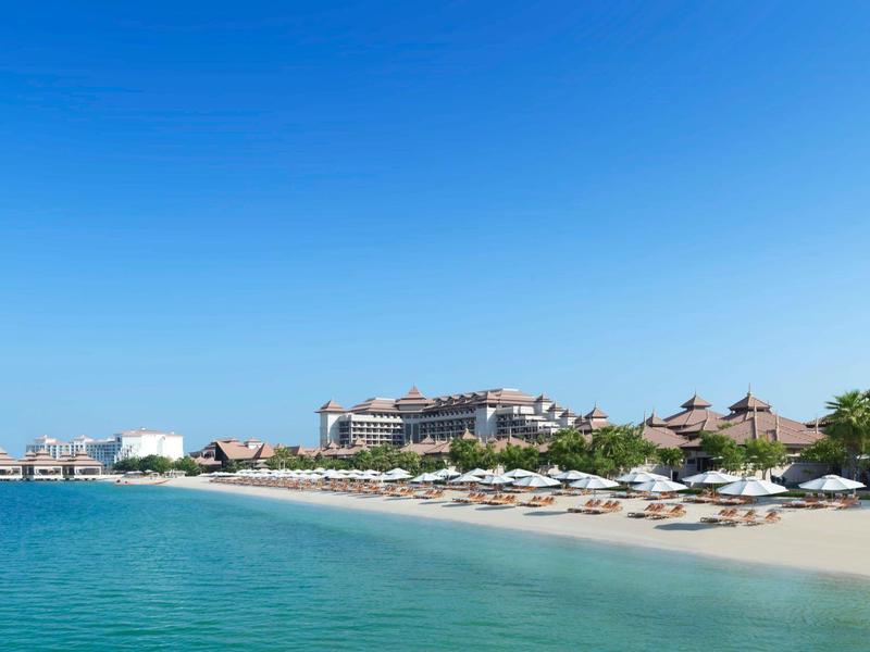 Strand mit weißem Sand, türkisfarbenem Meer und Gebäudeensemble unter klarem blauem Himmel.