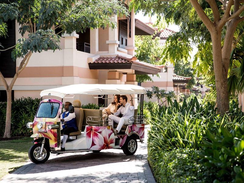 Weiße Rikscha mit Fahrgästen fährt auf einem von Bäumen gesäumten Weg vor einem Haus.