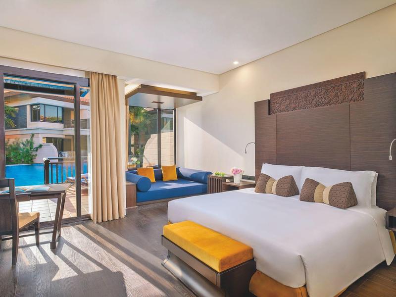 Helles Hotelzimmer mit großem Bett, gelber Sitzbank, Sofa und Glasfenster zum Außenbereich mit Pool.