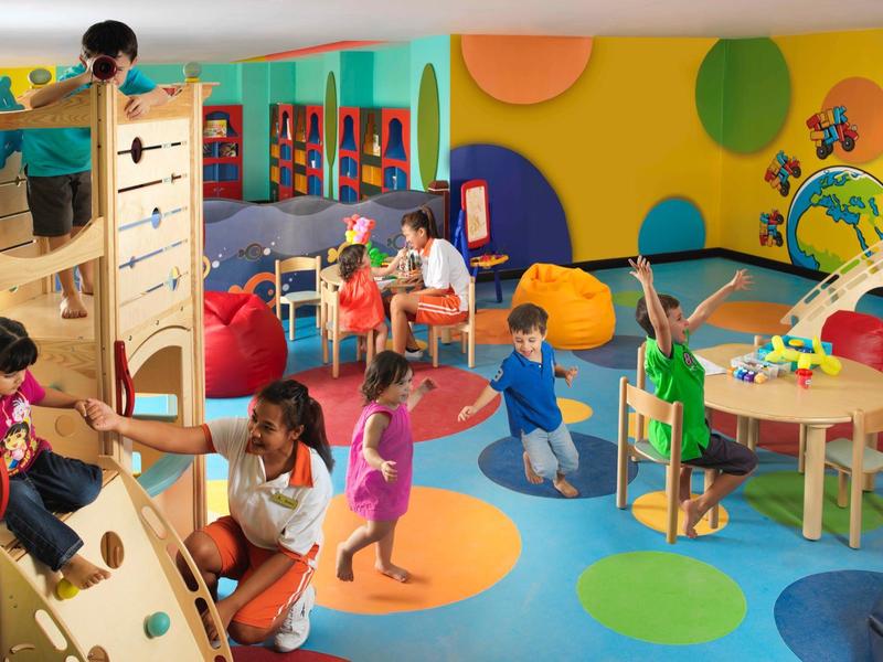Bunte Kinderspielfläche mit Kletterwand, Bällen, Tischen und spielenden Kindern und Betreuern.
