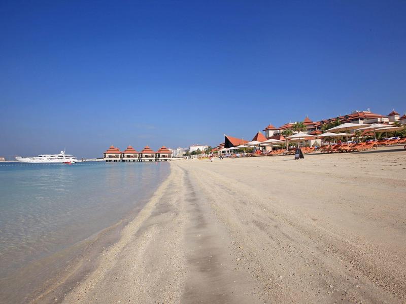 Breiter Sandstrand mit klarem Meer und Häusern am Hang unter blauem Himmel.