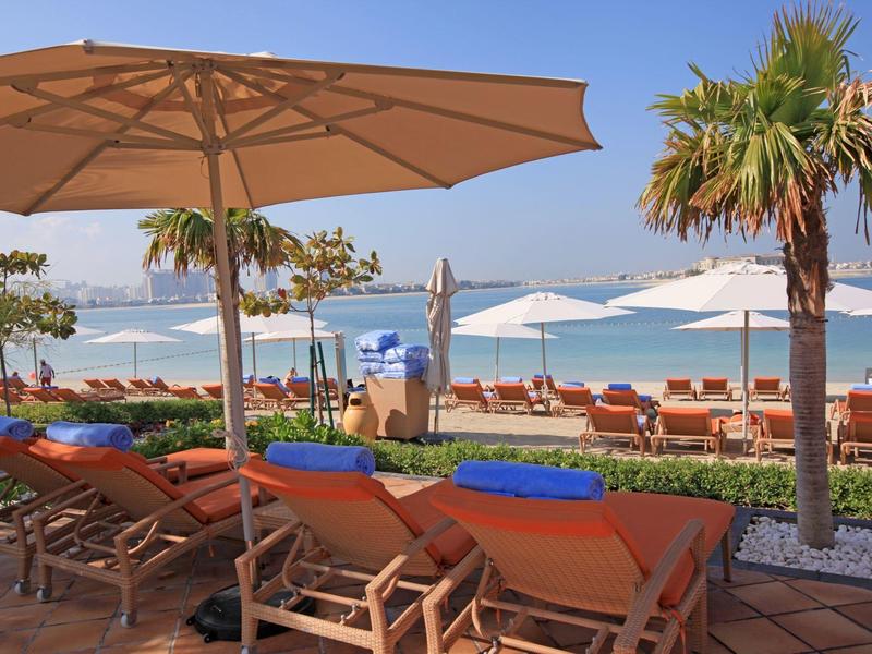 Strand mit Liegestühlen unter Sonnenschirmen, Palmen und blauem Himmel am ruhigen Meer.
