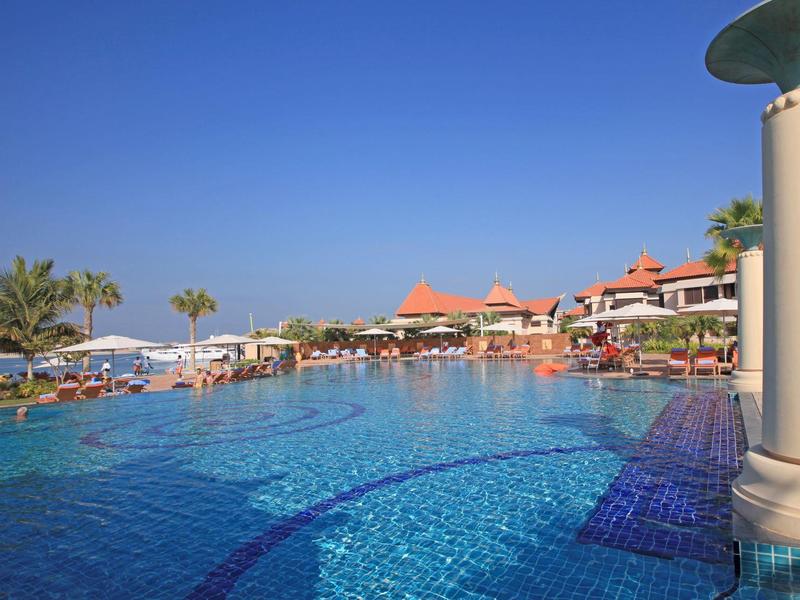 Großer blauer Pool mit Sonnenschirmen, Liegen und mediterranen Gebäuden unter klarem Himmel.