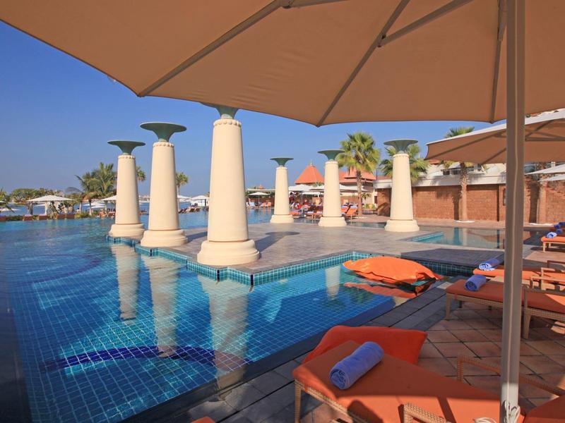 Poolbereich mit Säulen, Sonnenschirmen und Liegestühlen mit orangenen Handtüchern, blauer Himmel.