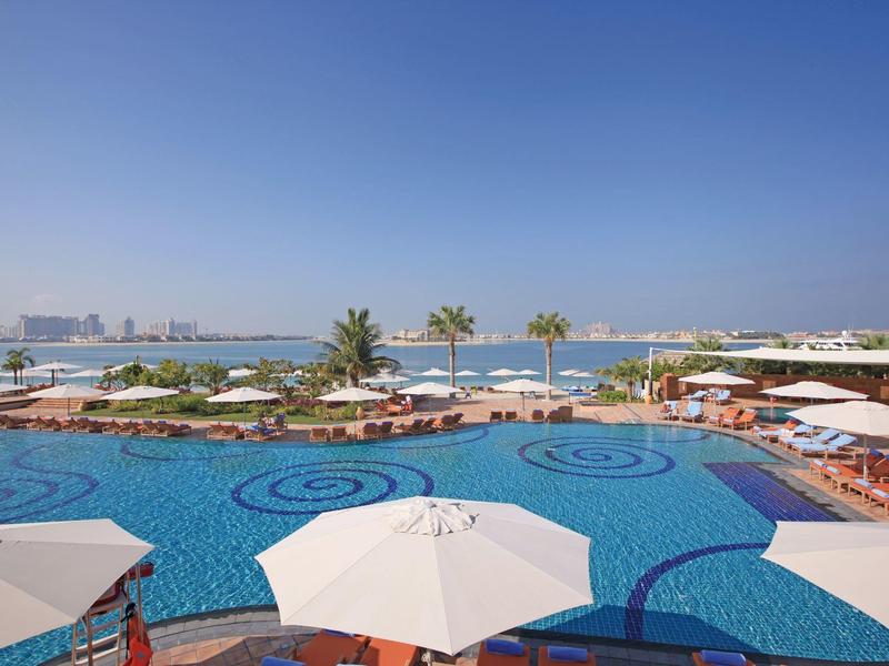 Großer blauer Pool mit weißen Sonnenschirmen und Palmen am Meer bei klarem Himmel.