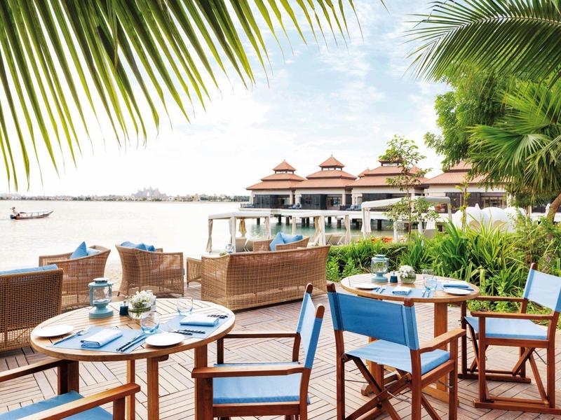 Strandrestaurant mit Holzmöbeln, blauer Bestuhlung, Palmen und Blick aufs Meer bei klarem Himmel.