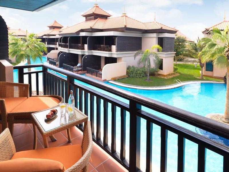 Balkon mit Stühlen und Tisch, Blick auf tropischen Pool und moderne Hotelgebäude bei Sonnenschein.