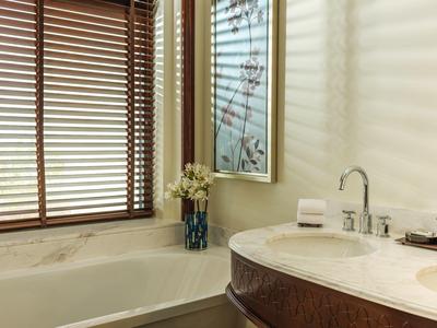 Helles Badezimmer mit weißer Badewanne, Holzjalousien, rundem Waschbecken und Vase mit Blumen.