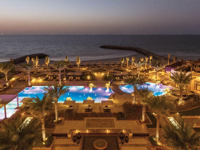 Blick auf beleuchteten Poolbereich und Palmen am Abend mit Meer und Hafen im Hintergrund.