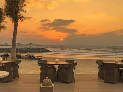 Strandrestaurant bei Sonnenuntergang mit Tischen, Stühlen und dekorativen Palmen am Holzdeck.