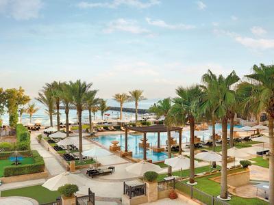 Luxuriöse Hotelanlage mit Swimmingpool, Palmen und Liegestühlen am Meer bei klarem Himmel.