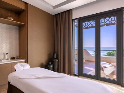 Helles Behandlungszimmer mit Massageliege, großem Fenster und Blick auf Balkon und Meer.
