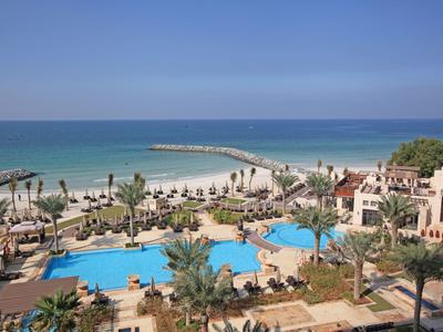 Blick auf einen Poolbereich mit Palmen, Strand und Meer im Hintergrund bei klarem Himmel.