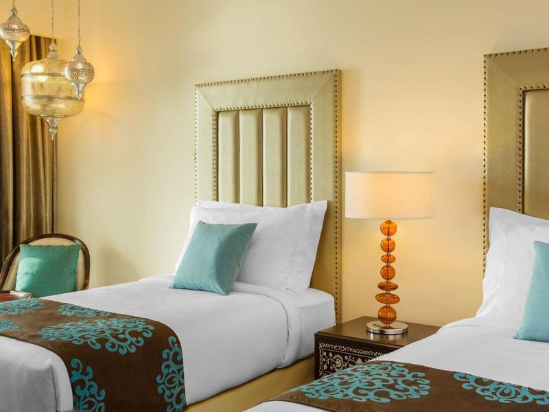 Zwei Einzelbetten mit weißen Laken, türkisfarbenen Kissen und braunem Muster, daneben Nachttisch und Stuhl.