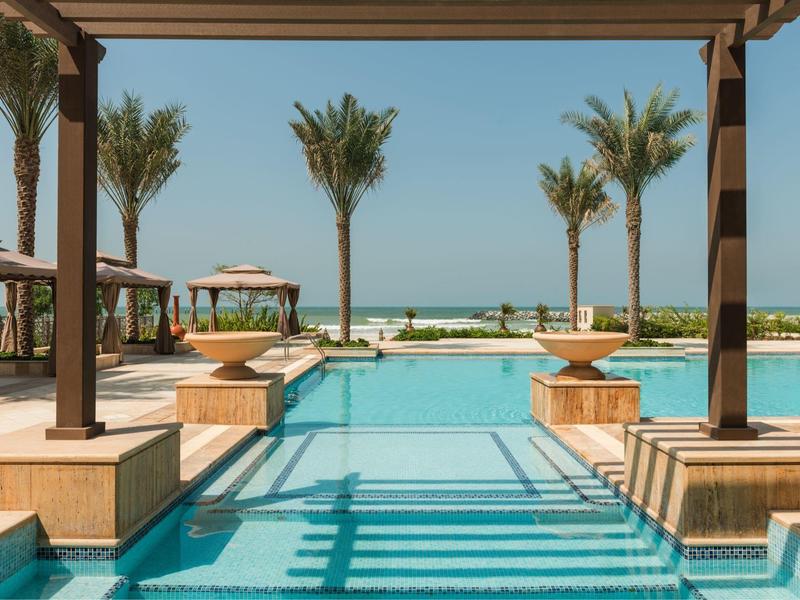 Moderner Pool mit Holzliegen, Palmen und Blick auf das Meer bei klarem Himmel.