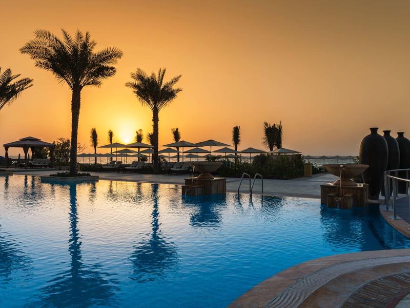 Pool mit Palmen und Liegen, Sonnenuntergang am Meer im Hintergrund, ruhige Wasseroberfläche.