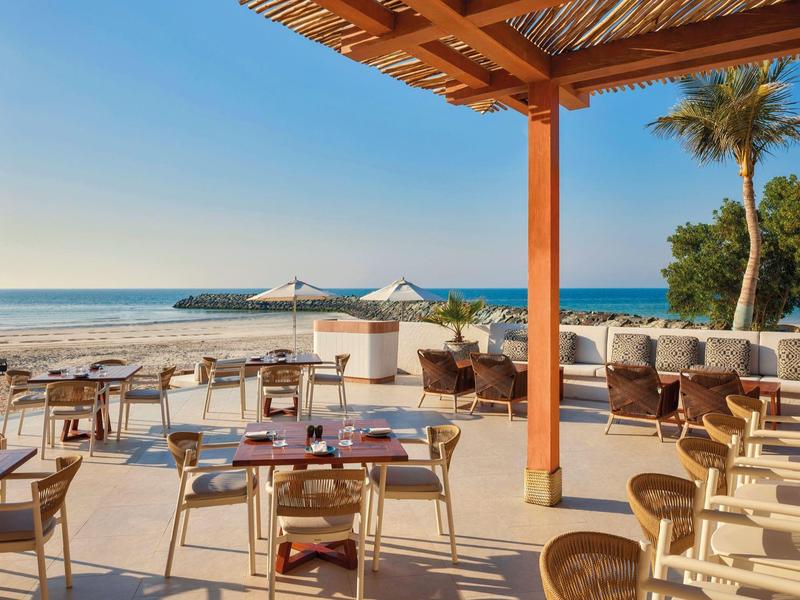 Offenes Strandrestaurant mit Holzmöbeln, Sonnenschirmen und Blick auf ruhiges blaues Meer.