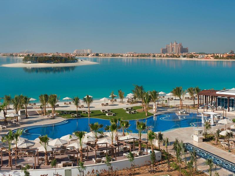 Großer Pool mit Palmen, Liegestühlen und Blick auf türkisblaues Meer und Inseln im Hintergrund.