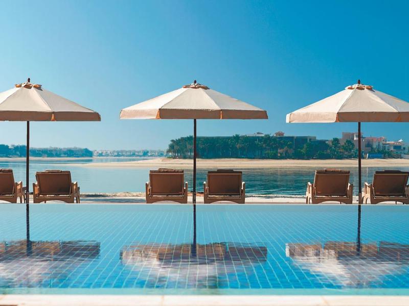 Drei Sonnenschirme und Liegestühle am blauem Infinity-Pool mit Meer und Insel im Hintergrund.