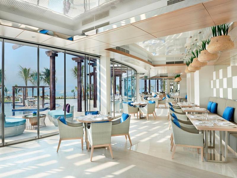 Moderner heller Innenraum mit großen Fenstern, weißen Möbeln, blauen Kissen und Meerblick.