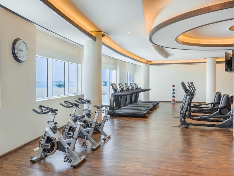 Moderner Fitnessraum mit Fahrradergometern, Laufbändern, hellen Fenstern und hölzernem Boden.