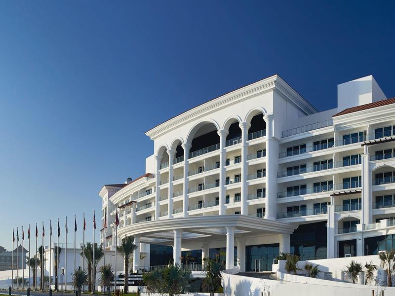 Großes weißes Hotelgebäude mit Rundbögen und vielen Balkonen vor klarem blauem Himmel.