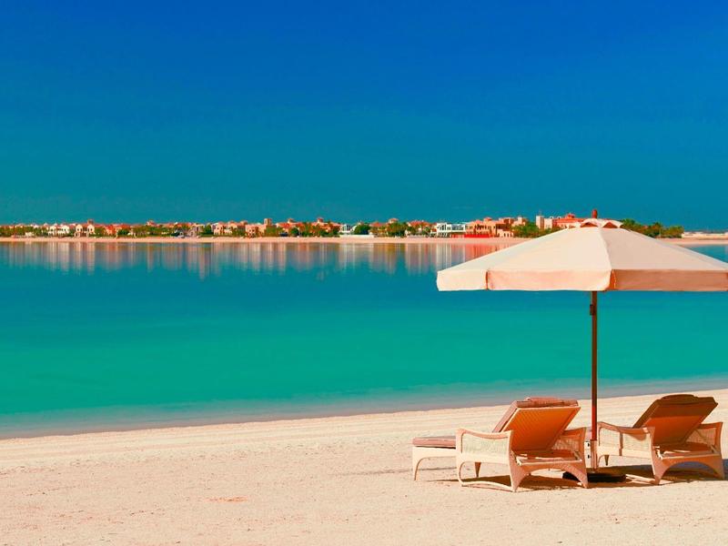 Strand mit weißem Sand, türkisfarbenem Wasser, zwei Liegestühlen und einem Sonnenschirm bei klarem Himmel.