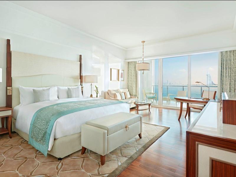 Helles Schlafzimmer mit großem Bett, Sitzbank, Holzmöbeln und Meerblick durch große Fenster.