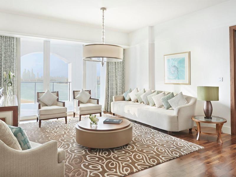 Helles Wohnzimmer mit Holzfußboden, beigen Sofas, rundem Teppich und großem Fenster mit Vorhängen.