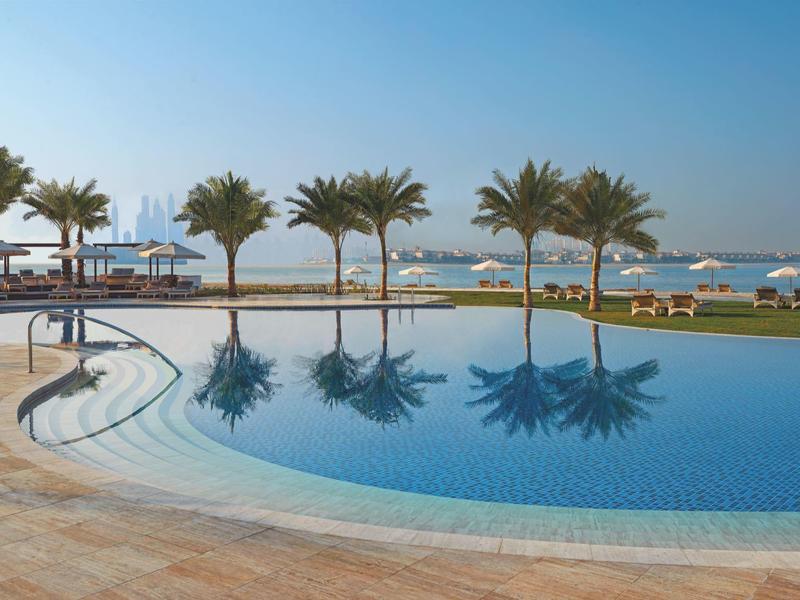Runder Pool mit spiegelglattem Wasser, Palmen und Sonnenschirmen am Strand bei klarem Himmel.