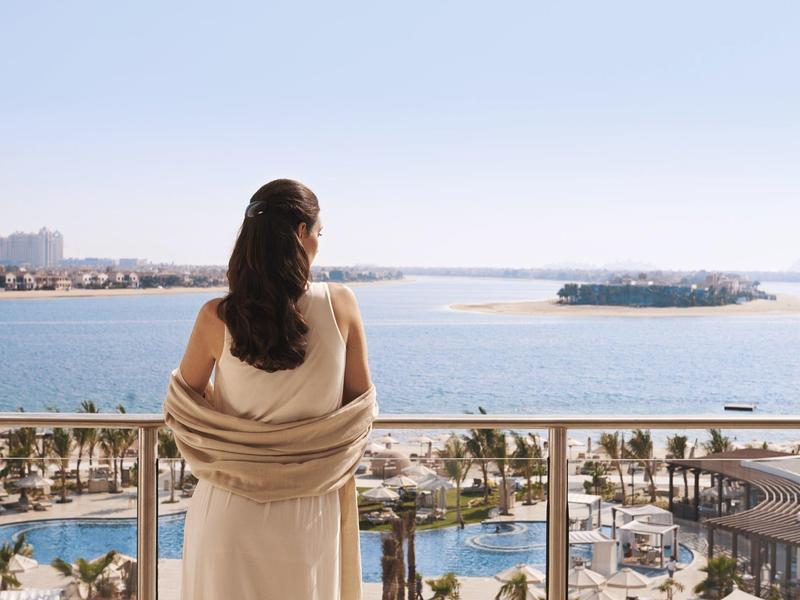 Frau in hellbeigem Kleid mit braunem Haar steht am Balkon und blickt auf Meer und Strand.