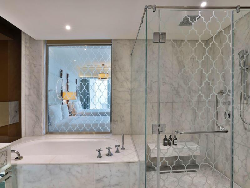 Modernes Badezimmer mit Glasdusche, hellgrauen Fliesen und Blick in ein helles Schlafzimmer.