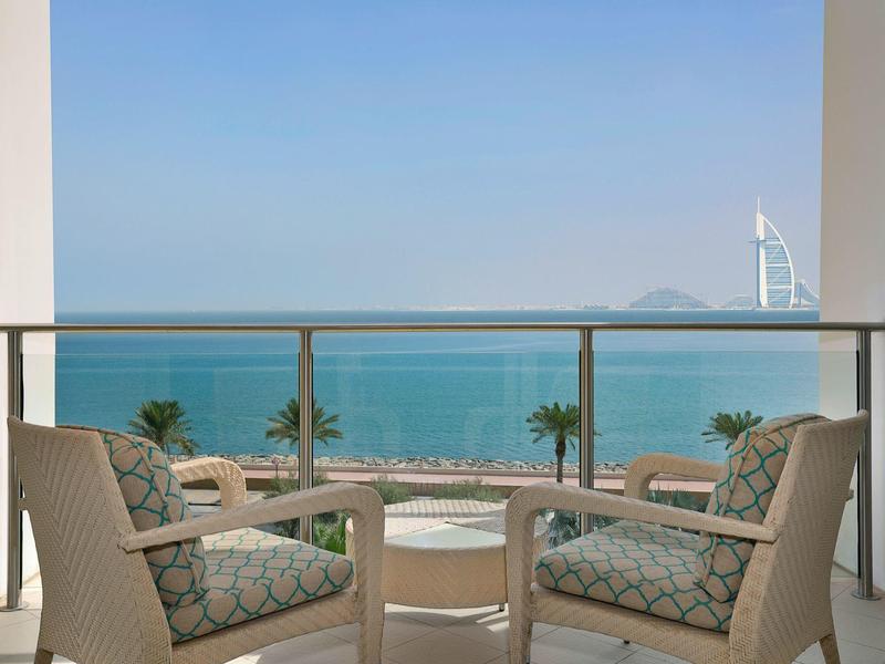Balkon mit zwei Sesseln und Tisch, Blick auf Meer, Strand, Palmen und modernes Hochhaus am Horizont.
