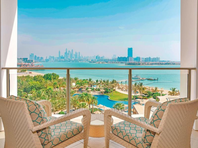 Zwei Stühle auf Balkon mit Glasgeländer, Blick auf Pool, Palmen und Stadt mit Hochhäusern am Strand.