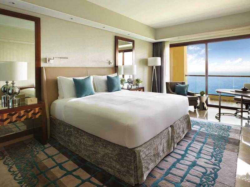 Modernes Schlafzimmer mit großem Bett, Teppich, Spiegel, Sesseln und Meerblick durch Fenster.