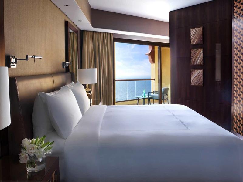 Modernes Hotelzimmer mit großem Bett, Meerblick durch Fenster, Holzwänden und Vorhängen in warmen Tönen.